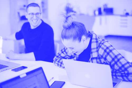 Zwei Personen sitzen an einem Tisch und arbeiten an Laptops. Die Person links trägt eine Brille, lächelt und blickt in Richtung der Person rechts, die ein kariertes Hemd trägt und lachend nach unten schaut. Die Szene ist mit einer blauen Farbüberlagerung versehen.