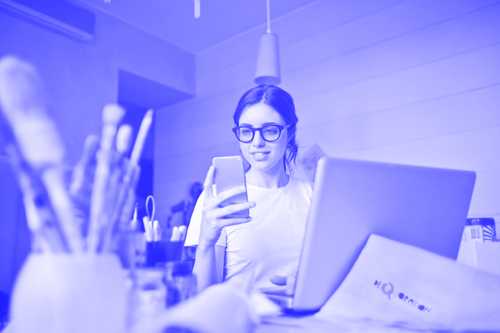 Eine Person mit Brille sitzt an einem Schreibtisch, hält ein Smartphone in der Hand und schaut darauf, während sie von einem Laptop, Papieren und verschiedenen Kunstutensilien umgeben ist. Das Bild hat einen blau getönten Filter.