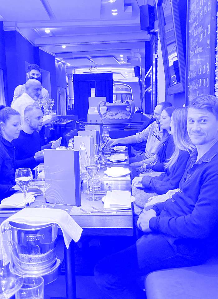 Eine Gruppe von Menschen sitzt in einem Restaurant um einen langen, schmalen Tisch. Einige unterhalten sich, andere blicken in die Kamera. Weingläser, Speisekarten und eine Champagnerflasche kühlen in einem Eimer auf dem Tisch. Die Atmosphäre ist locker und gemütlich.