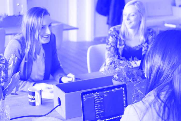 Eine Gruppe von vier Frauen sitzt in einem hellen Büro um einen Holztisch und diskutiert. Eine Frau benutzt einen Laptop mit sichtbarem Bildschirm und auf dem Tisch steht eine Tasse mit der Aufschrift „Boss Woman“. Auf dem Tisch liegen verstreut Pflanzen und Papiere.
